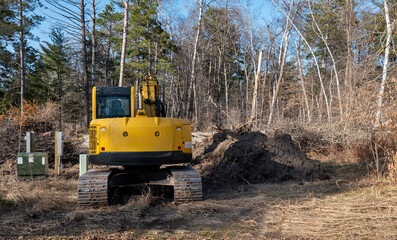 Yellow hydraulic backhoe excavator with tracks is parked near utility boxes at a construction site, with trees in the distance.