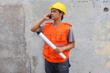 Obraz na płótnie Canvas Young Asian engineer in yellow safety helmet using mobile phone to make a call while holding white cardboard or blueprint paper standing near building wall