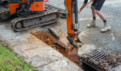 Excavator digging up dirt between cut concrete pavement. Man sweeping dirt with broom. Roadworks in...