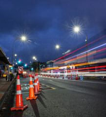 Orange traffic cones lining up the road. Bus light trails approaching the intersection. Roadworks...