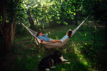 Two beautiful happy lesbian girls relax and hold hands in a hammock in the garden with their dog in the rays of the sun - obrazy, fototapety, plakaty