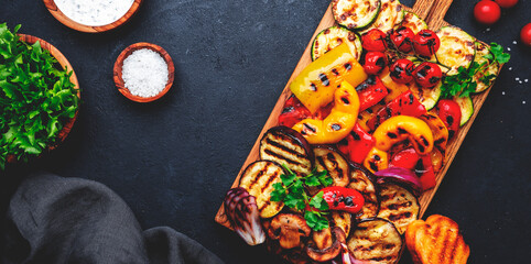 Grilled vegetables on rustic wooden cutting board,: colorful paprika, zucchini, eggplant, mushrooms, tomatoes and red onions with yogurt dressing, black stone table background, top view