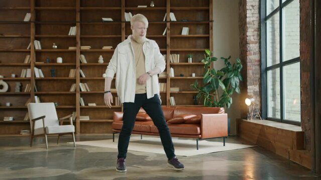 Happy young man dancing in loft style living room. Man wearing hipster casual clothes, jumping and moving hands around in ecstatic moves. Blond bearded guy listening to music at home on sunny morning
