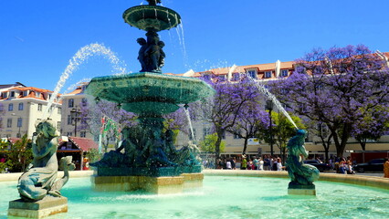 alter Brunnen mit Figuren und Wasserfontainen auf  Rossio Platz in Lissabon vor blauen...