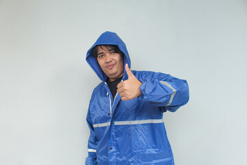 Asian young man with happy expression and thumbs up sign gesture in blue raincoat on gray background.