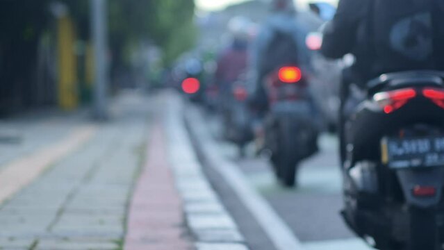 A blur image of Jakarta's urban traffic density dominated by motorcycles with a sidewalk on the side. Depicts high pollution, heat, traffic jams, fatigue from work, and inequality. 