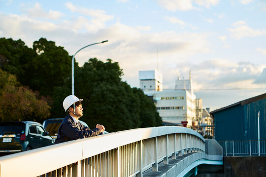 橋の上から遠くを見ている作業服とヘルメットの男性