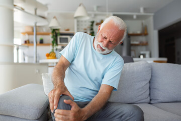 The older man is sitting on the couch at home, has pain in the knee joint, holding his leg,...