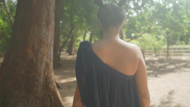 The camera follows a girl in a dress with partially exposed back and hair tied in small ponytails. Walking among the trees on a sunny day. Sunrays streaming through objects.