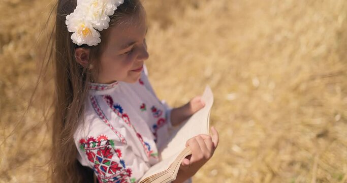 Girl in bulgarian embroidery ethnic folklore costume reading book on a harvesting wheat field, Bulgaria