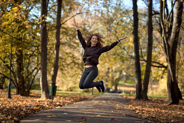 Wide shot of caucasian woman jumping in the park in the autumn   with arms outstretched - Powered by Adobe