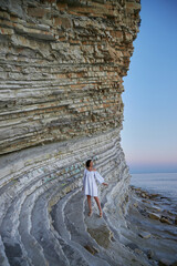 woman in white dress leaning on rock ledge, in the style of creased crinkled wrinkled, playful body...