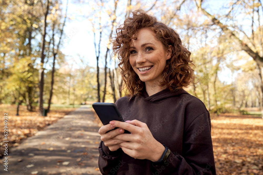 Wall mural Caucasian woman browsing mobile phone and looking away in the autumn park