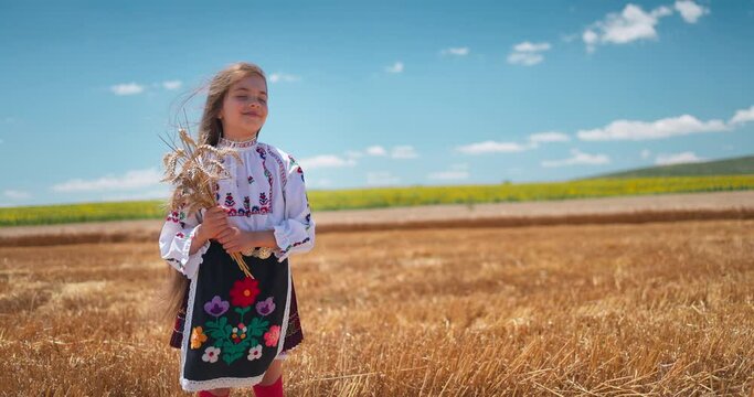 Girl in traditional ethnic folklore costume with Bulgarian embroidery standing on a harvest golden wheat field, Bulgaria agriculture 4K video
