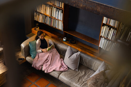 Biracial woman sitting on sofa reading book in living room, copy space