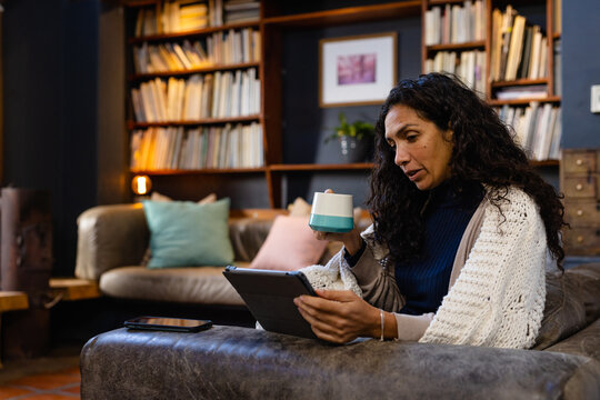 Biracial woman sitting on sofa holding mug and using tablet in living room