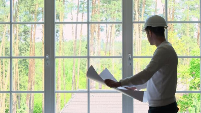 Architect standing by French windows learns residence building plan. Builder wearing white helmet preparing to renovate summer house