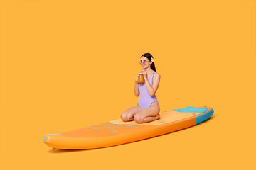 Happy woman with refreshing drink resting on SUP board against orange background