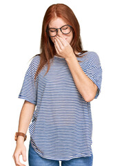 Young read head woman wearing casual clothes and glasses smelling something stinky and disgusting, intolerable smell, holding breath with fingers on nose. bad smell - obrazy, fototapety, plakaty