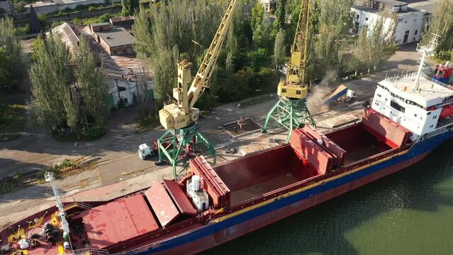 Grain transshipment from container to ship cargo holds with dual scoop clamshell grabs on gantry crane in sea port. Wheat shipment from dump trucks with water transport. Commodities transportation.