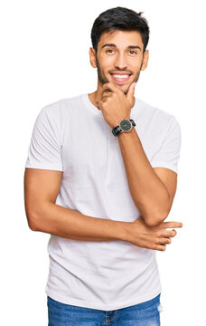 Young handsome man wearing casual white tshirt looking confident at the camera smiling with crossed arms and hand raised on chin. thinking positive.