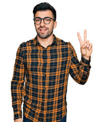 Hispanic man with beard wearing casual clothes and glasses smiling with happy face winking at the camera doing victory sign. number two.