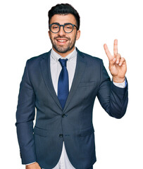 Hispanic man with beard wearing business suit and tie smiling with happy face winking at the camera doing victory sign. number two.