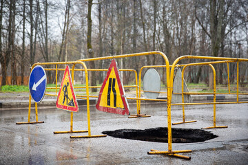 Large pit on the road with a fence and warning signs. Heavy rains have washed away the ground. Subsidence of soil. Collapse