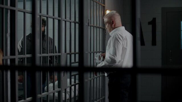 Prison guard puts handcuffs on criminal through metal bars, opens jail cell. Depressed prisoner goes to court session. Inmates serve imprisonment terms in detention center or correctional facility.
