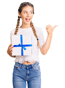 Beautiful caucasian woman with blonde hair holding gift pointing thumb up to the side smiling happy with open mouth