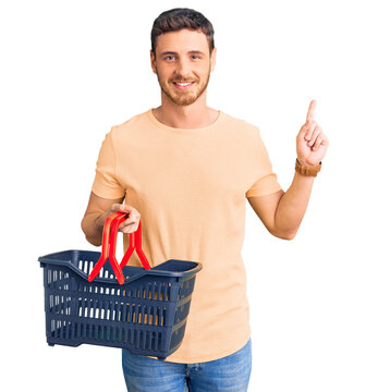 Handsome young man with bear holding supermarket shopping basket surprised with an idea or question pointing finger with happy face, number one
