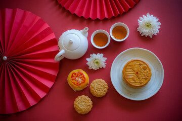 Chinese Mid-Autumn Festival concept made from mooncakes, tea decorated with Chrysanthemum blossom, rabbit and red paper fans on red background.