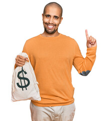 Hispanic adult man holding dollars bag surprised with an idea or question pointing finger with...
