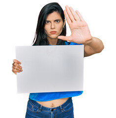 Young hispanic woman holding blank empty banner with open hand doing stop sign with serious and confident expression, defense gesture