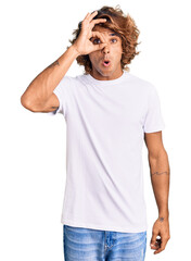 Young hispanic man wearing casual white tshirt doing ok gesture shocked with surprised face, eye looking through fingers. unbelieving expression.