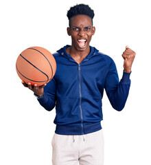 Young african american man holding basketball ball screaming proud, celebrating victory and success very excited with raised arms