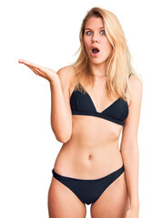 Young beautiful blonde woman wearing bikini surprised pointing with hand finger to the side, open mouth amazed expression.