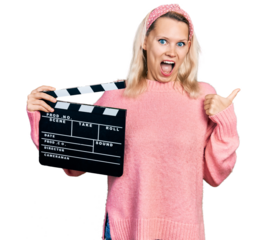 Young caucasian woman holding video film clapboard pointing thumb up to the side smiling happy with open mouth