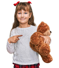 Little caucasian girl kid hugging teddy bear stuffed animal smiling happy pointing with hand and finger