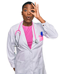 Young african american woman wearing doctor uniform and stethoscope doing ok gesture shocked with surprised face, eye looking through fingers. unbelieving expression.