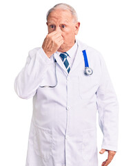 Senior handsome grey-haired man wearing doctor coat and stethoscope smelling something stinky and disgusting, intolerable smell, holding breath with fingers on nose. bad smell