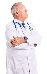 Senior handsome grey-haired man wearing doctor coat and stethoscope looking to the side with arms crossed convinced and confident