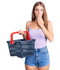 Young beautiful blonde woman holding supermarket shopping basket covering mouth with hand, shocked and afraid for mistake. surprised expression