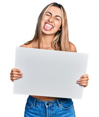 Hispanic young woman holding blank empty banner sticking tongue out happy with funny expression.