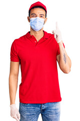 Young handsome hispanic man wearing delivery uniform and medical mask pointing finger up with successful idea. exited and happy. number one.