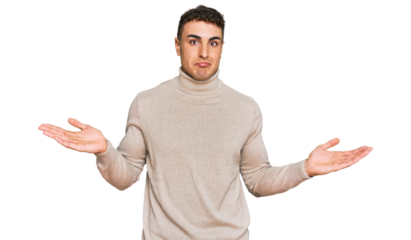 Hispanic young man wearing casual turtleneck sweater clueless and confused expression with arms and hands raised. doubt concept.