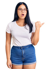 Young beautiful asian girl wearing casual clothes and glasses surprised pointing with hand finger to the side, open mouth amazed expression.