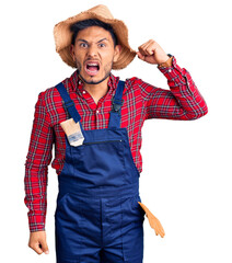 Handsome latin american young man weaing handyman uniform angry and mad raising fist frustrated and...