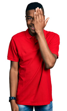 Young african american man wearing casual red t shirt covering one eye with hand, confident smile on face and surprise emotion.