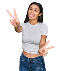 Young hispanic girl wearing casual white t shirt smiling with tongue out showing fingers of both hands doing victory sign. number two.